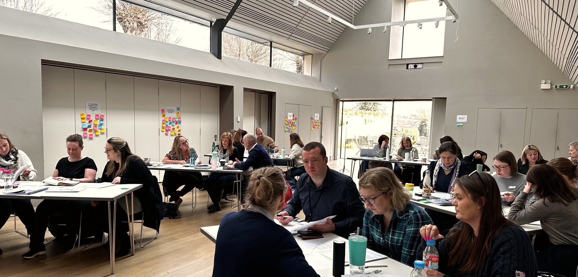 Leaders on an i3 Leadership Programme cohort working in small groups around tables in a bright hall.